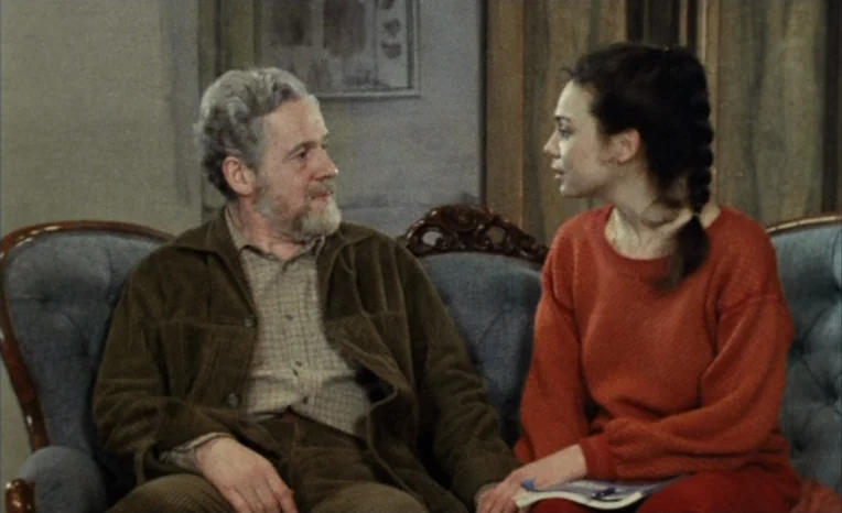 After The Rehearsal (1984) movie still: medium — A domestic space features a tufted blue sofa against a plain wall. An older man and a young woman…