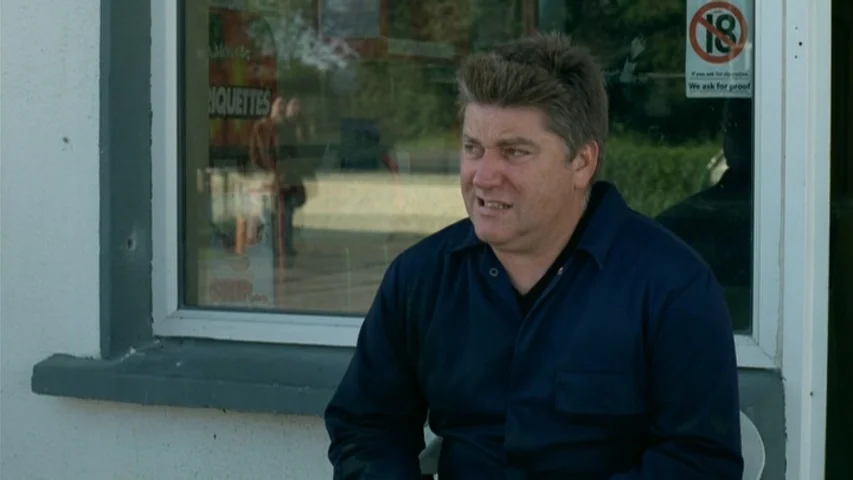 Garage (2007) movie still: medium — A man sits outside a store with an eighteen-plus sign visible on the window. He is engaged in conversation with…