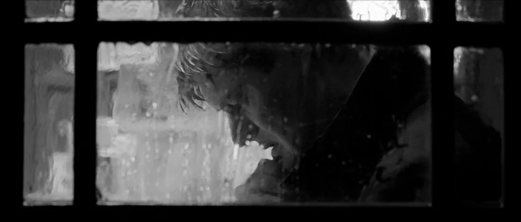 Control movie still: closeup — A dark, wet window pane frame obscures the view of a rainy night exterior. A man in a dark coat stands outside…