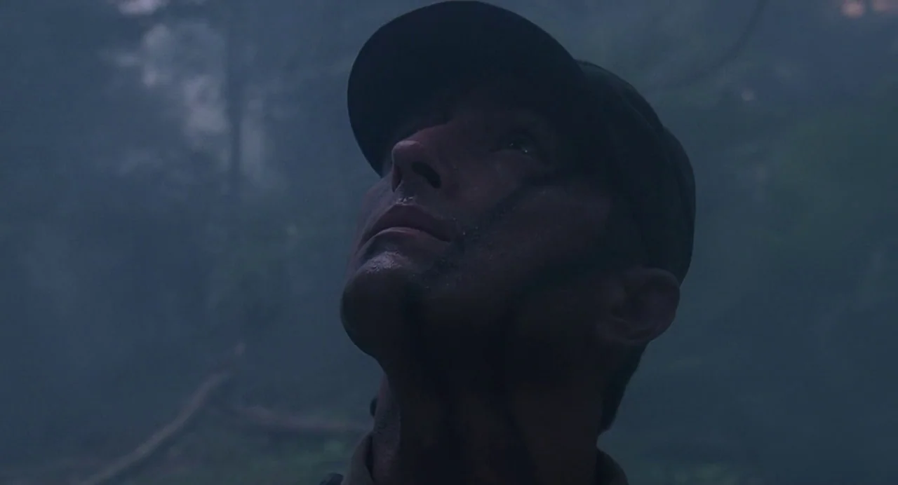 Predator (1987) movie still: closeup — A dense, misty jungle environment fills the frame behind a man. The man looks directly upward while wearing a…