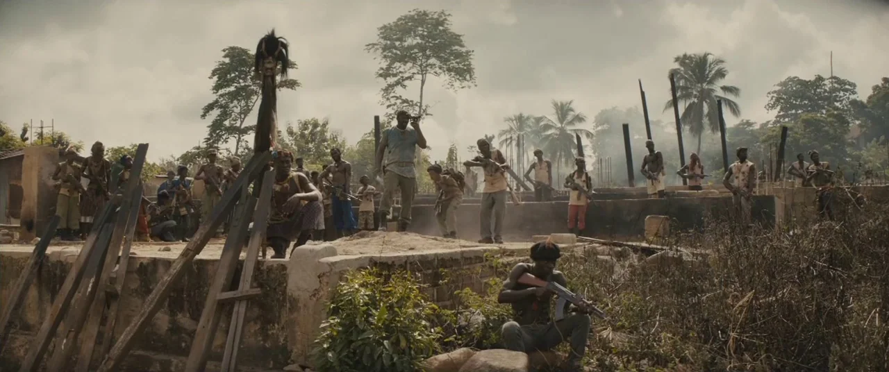 Beasts of No Nation (2015) movie still: wide — A group of armed men gather in the ruins of a concrete structure. Overcast sky looms over the leveled…