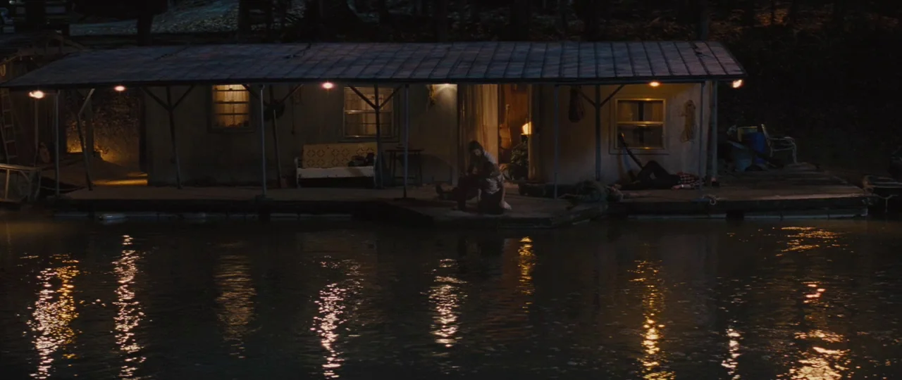 Mud (2012) movie still: wide — A small building sits on a wooden floating dock at night by the water. Several people are positioned on the planks in…