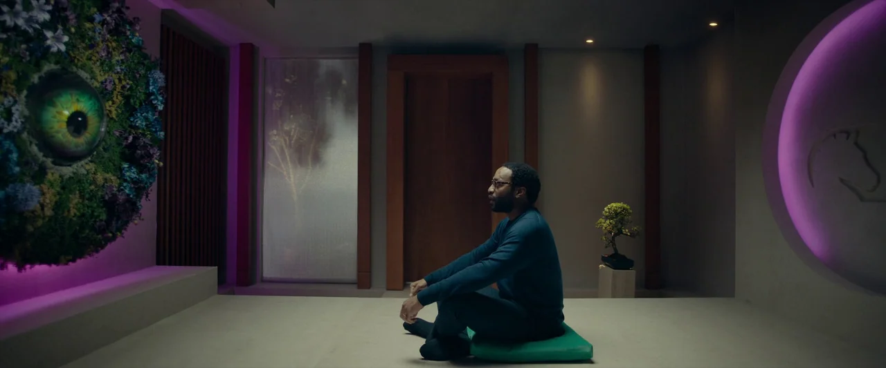 The Pod Generation (2023) movie still: wide — A minimalist room features smooth walls, a vertical light strip, and a large eye display. A man sits…