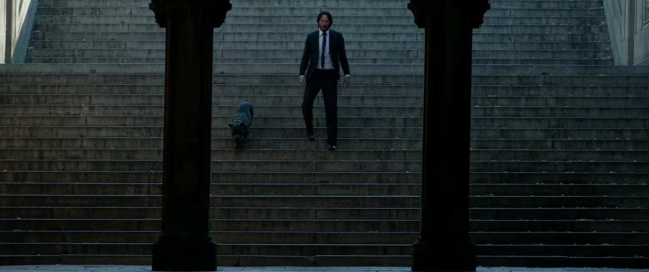 John Wick: Chapter 2 (2017) movie still: wide — A man and his dog descend a massive, dark set of stone steps outdoors. Large stone pillars flank the…