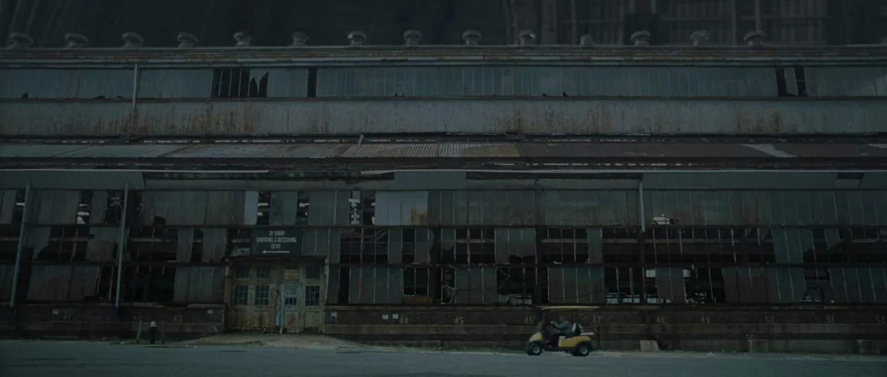 Synecdoche, New York (2008) movie still: wide — A long, rusted industrial building features rows of broken windows and metal siding panels. A small…
