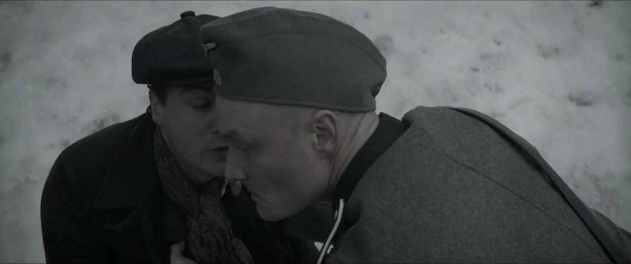 Bau, Artist at War (2025) movie still: medium — A snowy outdoor environment with two men interacting closely in the winter cold. One man in a…