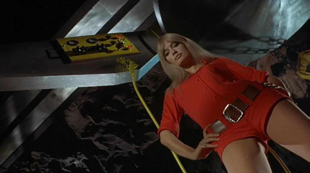 Danger Diabolik (1968) movie still: fullbody — A dark, rocky interior space features a futuristic yellow and silver control panel console. A blonde…
