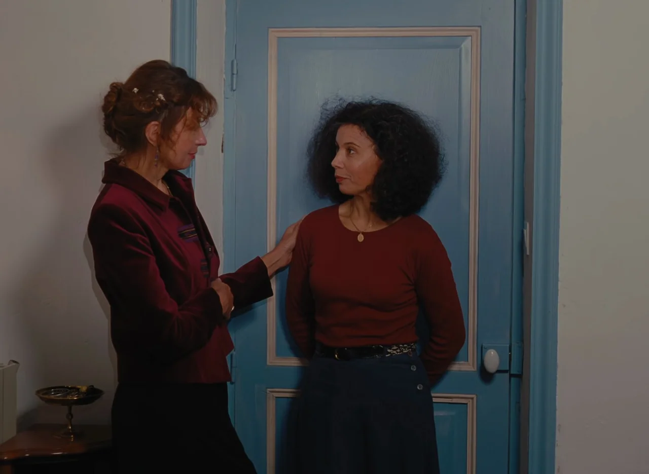 Autumn Tale (1998) movie still: medium — Two women stand before a blue-painted wooden door in a simple room. One woman places a hand on the other…