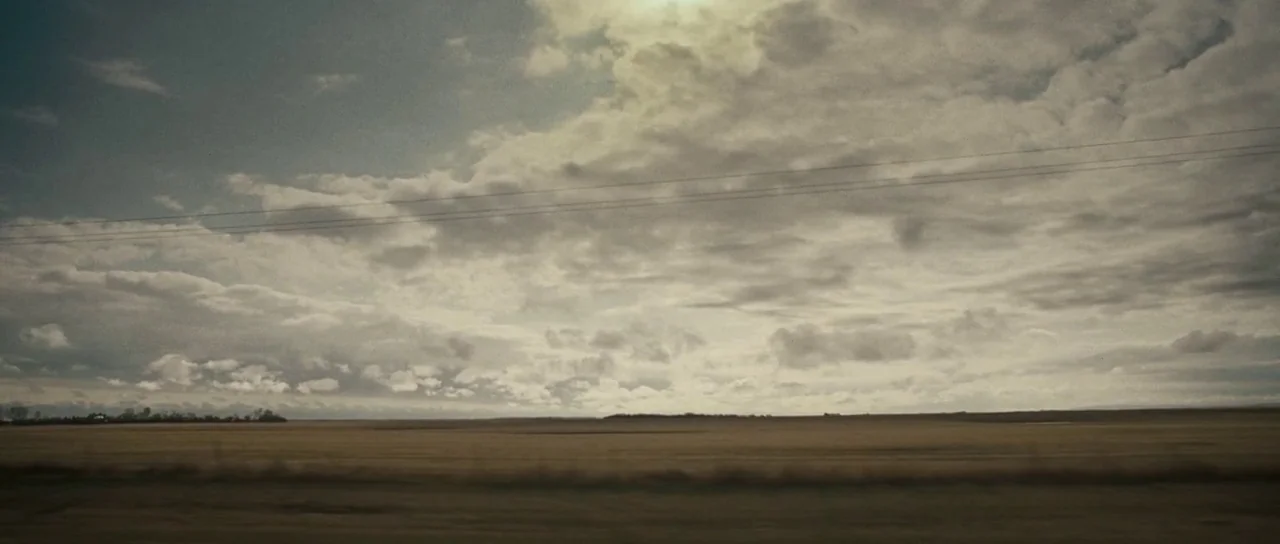 Surveillance (2008) movie still: establishing — A vast, flat agricultural field stretches to the horizon under a cloudy sky. Power lines are visible…