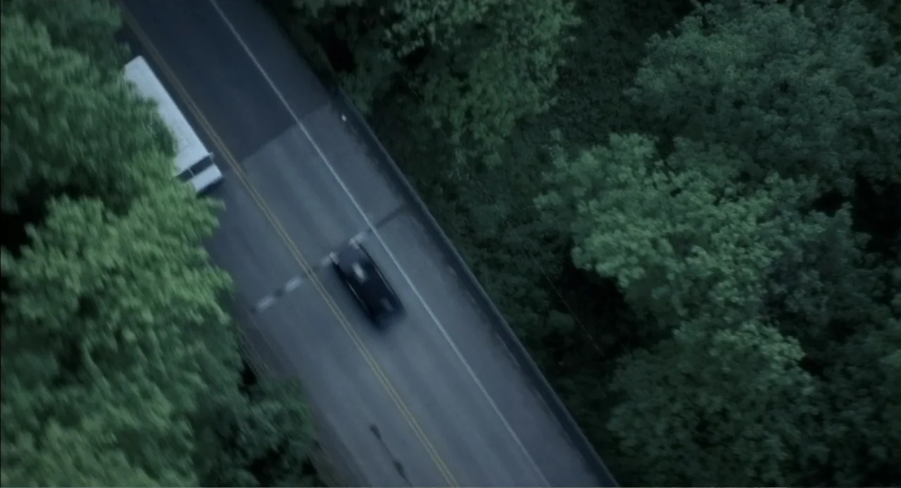 The Hunted (2003) movie still: establishing — An asphalt road cuts through a dense forest of mature green trees. A dark sedan travels along the road…