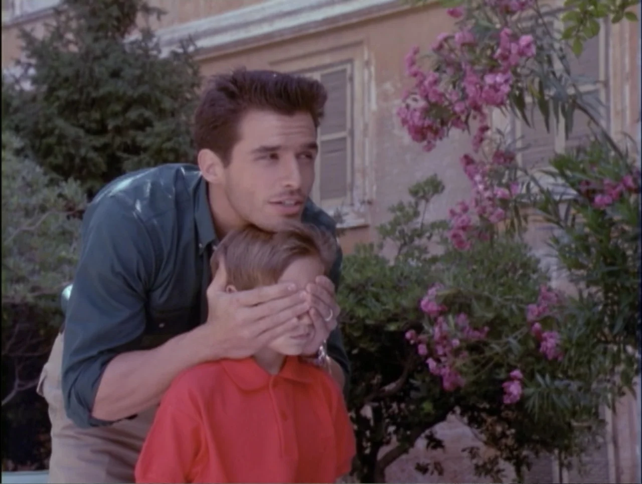 Code Name: Wolverine (1996) movie still: medium — An outdoor courtyard features an old stone building wall and lush pink blooming shrubs. An adult…