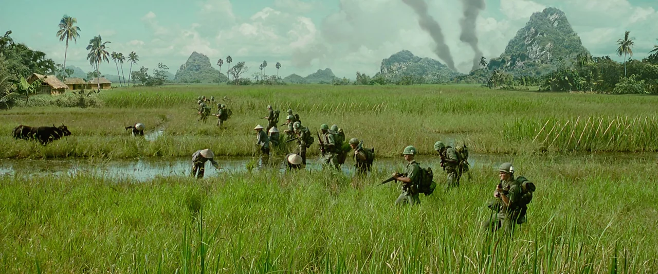 Forrest Gump (1994) movie still: wide — A green rice paddy landscape under a cloudy sky with distant smoking peaks. American soldiers march through…