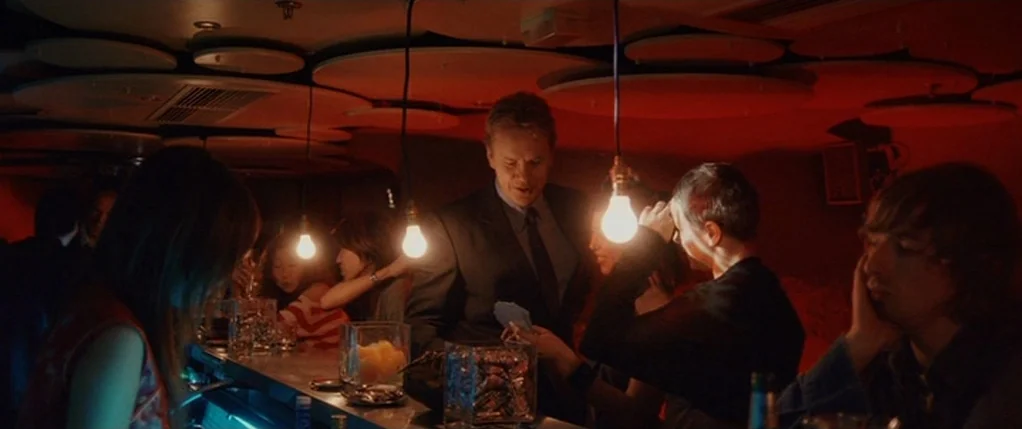 Code 46 movie still: medium — A dimly lit red bar features circular acoustic panels on the ceiling and hanging bulbs. People congregate around the…