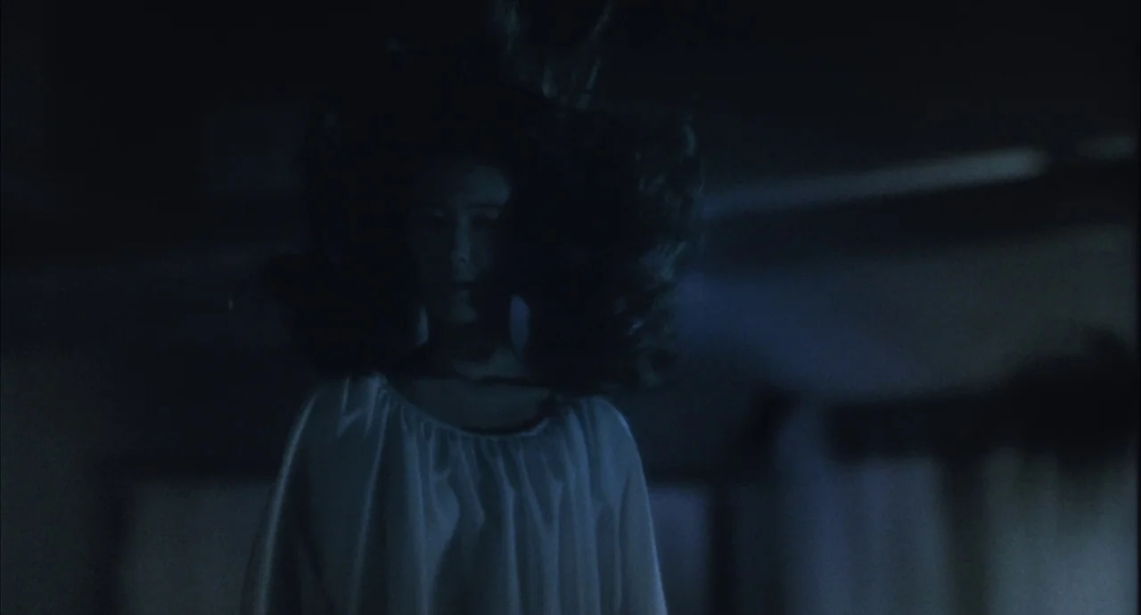 The Discarnates (1988) movie still: medium — A dark, dimly lit room is filled with shadow and indistinct architectural forms. A young girl in a…
