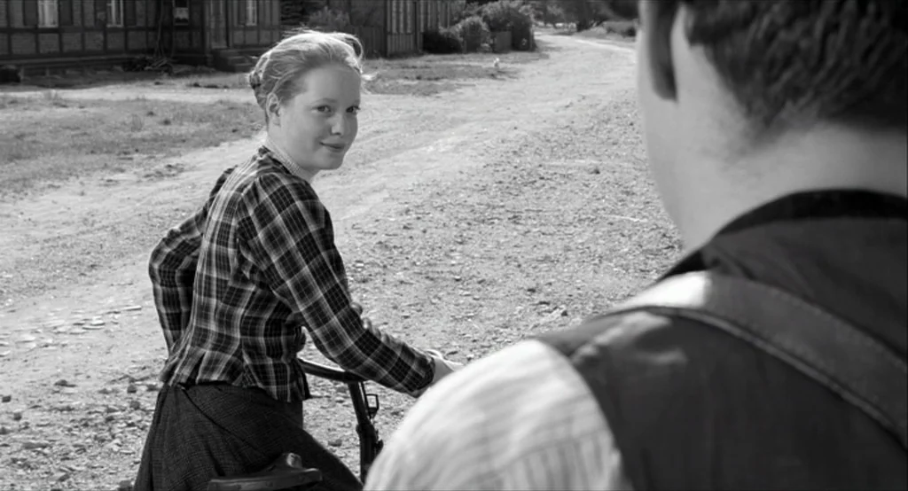 The White Ribbon (2009) movie still: medium — A rural dirt path runs between timber-framed buildings in a small village. A young woman on a bicycle…