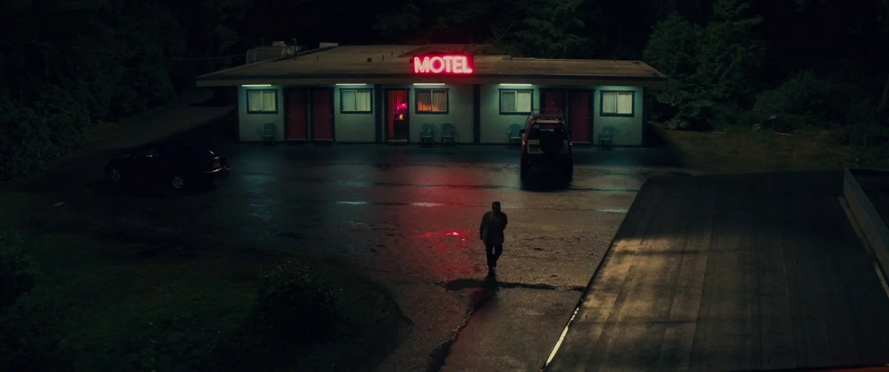 Come To Daddy (2019) movie still: wide — A dark, wet asphalt lot serves as the forecourt to a single-story motel building. A solitary man walks…