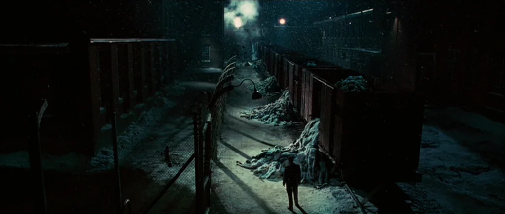 Shutter Island (2010) movie still: wide — A dark, snow-covered industrial railway yard at night with piles of bodies near train cars. A lone man in…