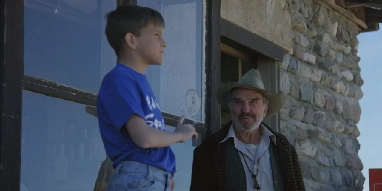 El sótano (2024) movie still: medium — An exterior of a rustic stone building features a young boy and an older man. The boy gestures toward the man…