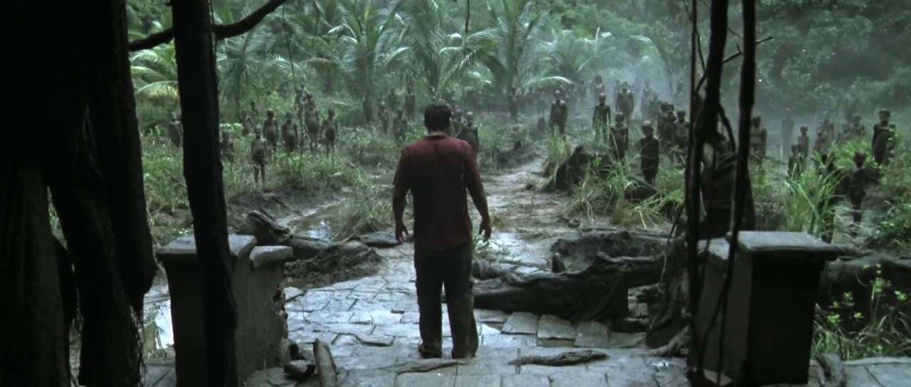 Vinyan (2008) movie still: wide — A man stands at the top of old stone steps leading into a humid jungle. A large group of people stands in the mud…