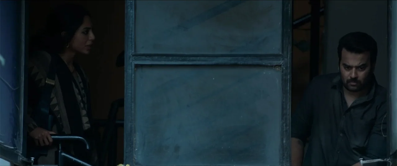 Cheekatilo (2026) movie still: medium — A dark, framed glass doorway separates a man and a woman in a dim space. They stand on opposite sides of the…