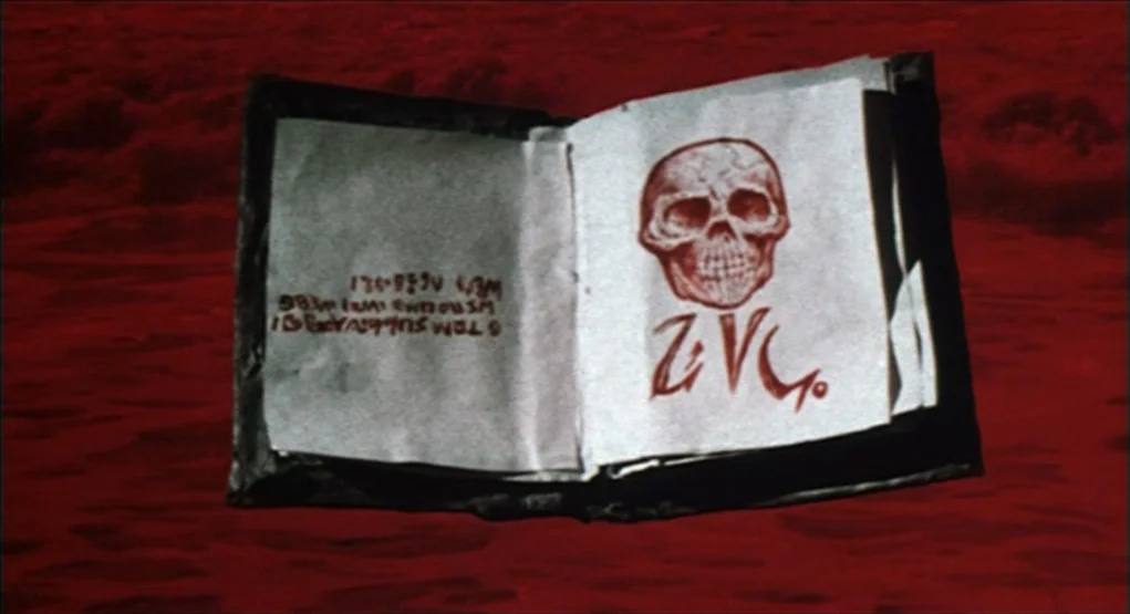 Evil Dead II (1987) movie still: closeup — An open book with a skull illustration rests on a dark red surface. The paper contains written text and a…