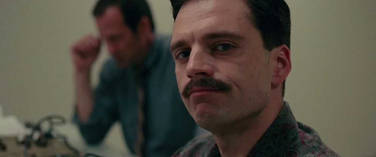 I, Tonya (2017) movie still: closeup — An austere room with plain light-colored walls and fluorescent ceiling lights. A man in a patterned shirt…