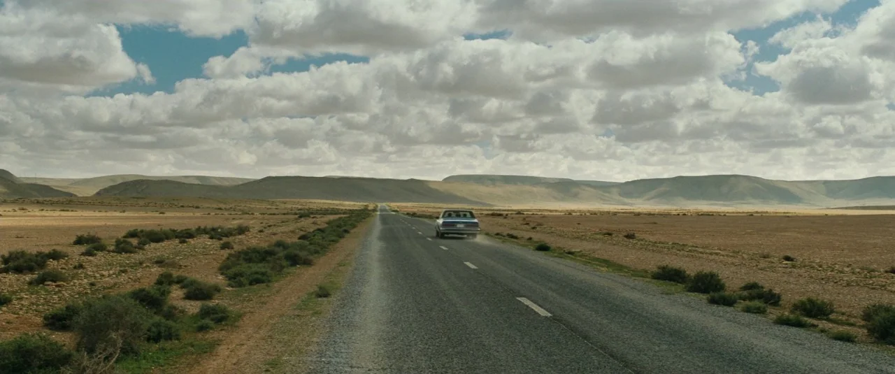 A Hologram for the King movie still: wide — An asphalt road cuts through a vast, arid desert landscape under a cloudy sky. A single sedan travels…