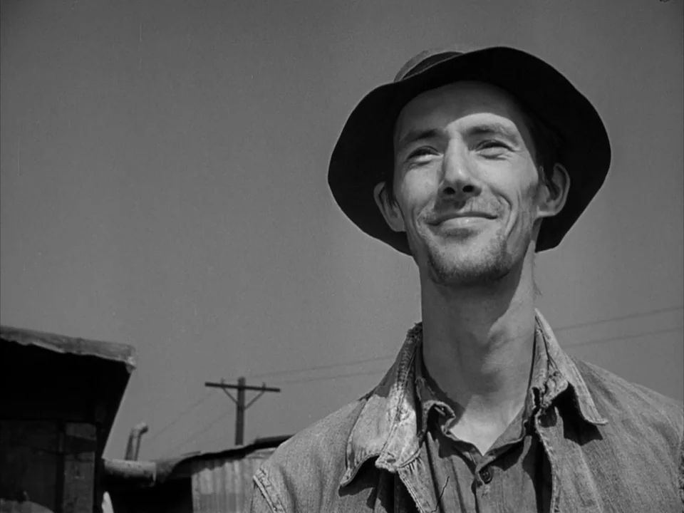The Grapes of Wrath (1940) movie still: closeup — A rural outdoor setting with a clear sky and distant telephone poles. A thin man in a hat looks…