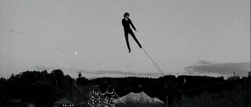 I’m Not There (2007) movie still: wide — An open landscape at twilight features a silhouette of a house with string lights. A man floats in the air,…
