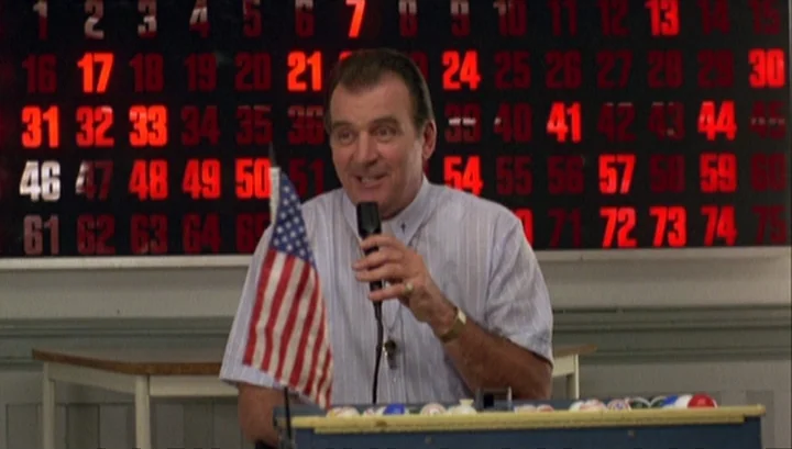 Highway 61 (1991) movie still: medium — A bingo hall interior features a dark wall with a large illuminated numeric grid. A man stands behind a…