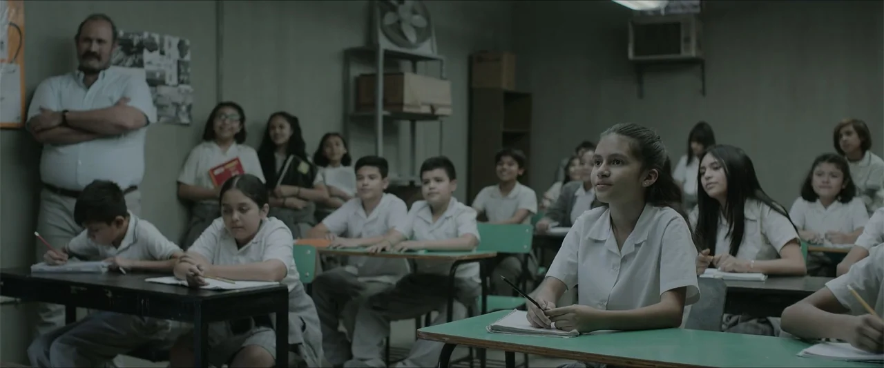 Radical (2023) movie still: medium — A classroom with grey walls and several rows of desks holds multiple seated students. A young girl in a white…