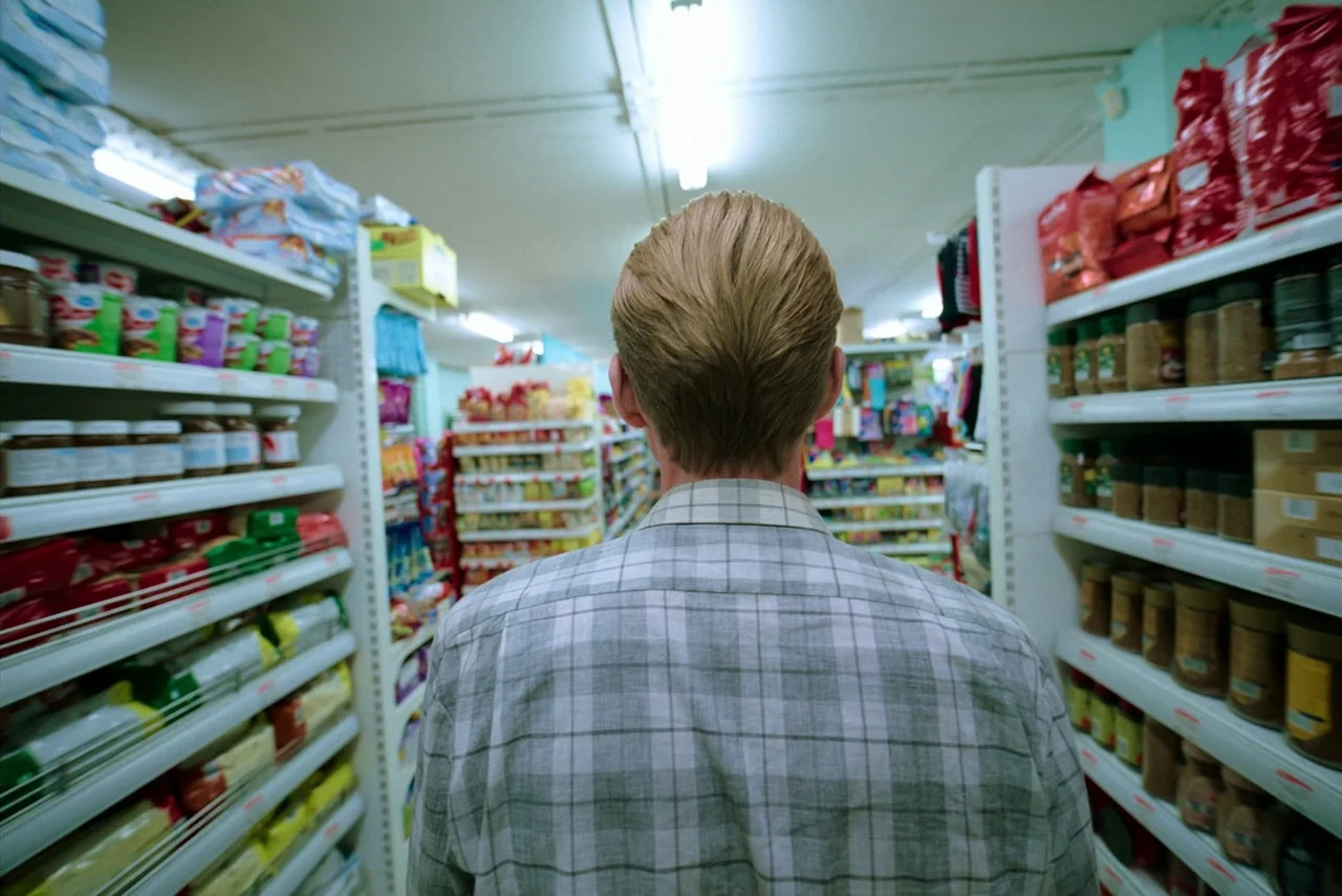 Barbarian (2022) movie still: medium — A narrow aisle in a grocery store is packed with shelves holding various food items. A person in a plaid…