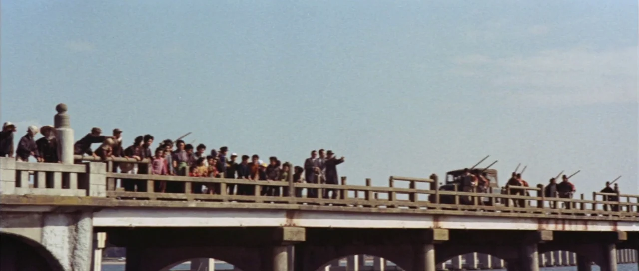 Mothra Vs Godzilla (1964) movie still: wide — A long concrete bridge stretches horizontally across the frame under a clear pale sky. Many people in…