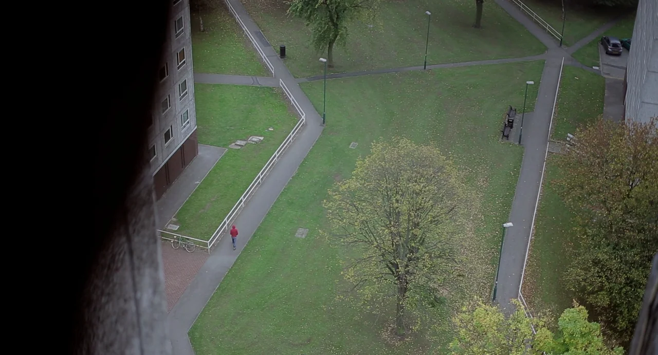Weekend (2011) movie still: wide — A sprawling public housing estate features concrete walkways winding through scattered trees and grass. A person…