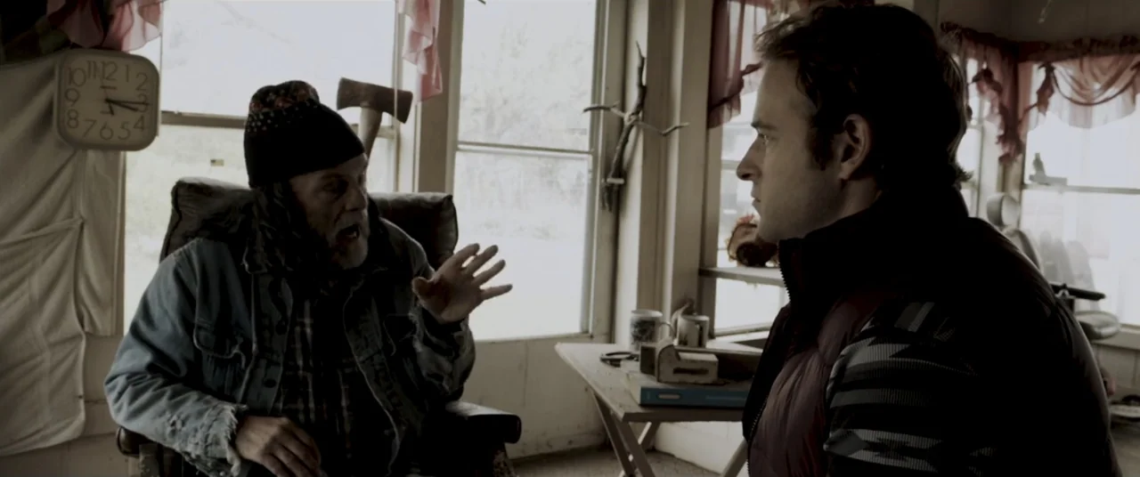 Amityville Cult (2021) movie still: medium — A cluttered, rustic interior features worn walls and light streaming through large windows. An older…