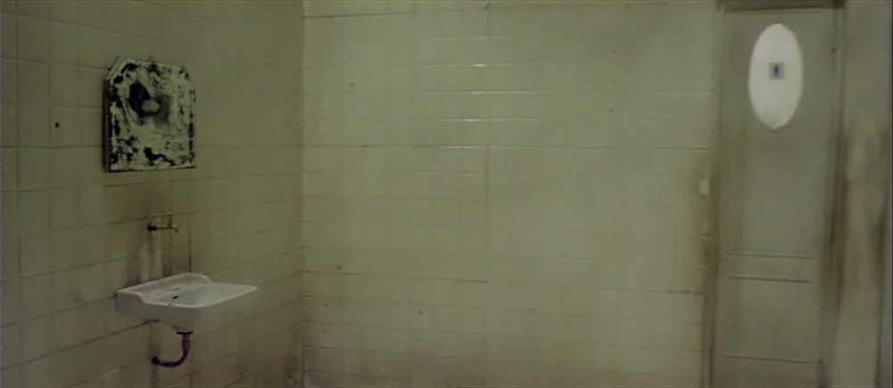 Deep Red (1975) movie still: establishing — A sparsely furnished public restroom features pale tiled walls and a single ceramic sink. A small,…