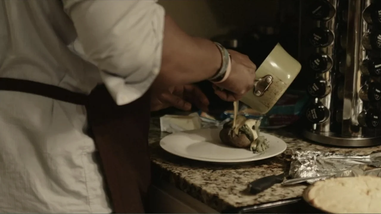 Death of a Child (2017) movie still: medium — A person stands at a granite kitchen counter preparing a meal. The individual pours sauce from a small…