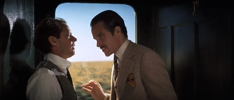The Fortune (1975) movie still: medium — Two men talk inside a dimly lit vintage train compartment during the daytime. A vast, blurred landscape…