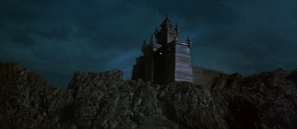 Dune (1984) movie still: establishing — A tall, wooden-walled castle structure sits atop a sharp, jagged rock formation at night. The sky is dark…