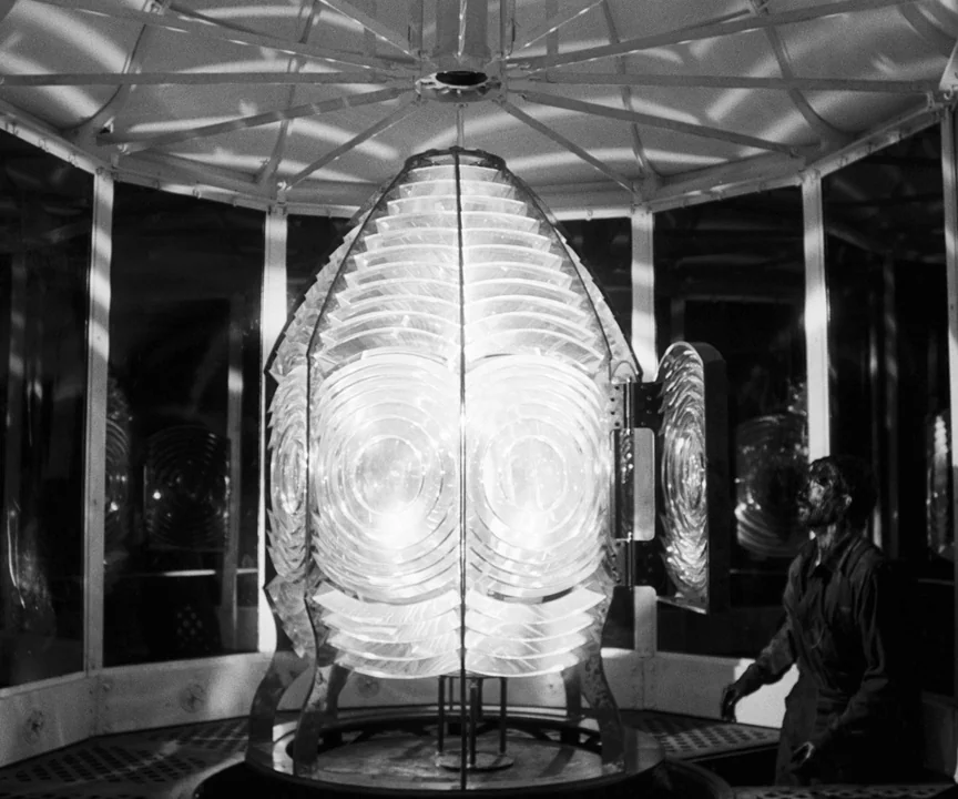 The Lighthouse (2019) movie still: wide — A glass-enclosed lantern room houses a massive, glowing multi-tiered fresnel lens at night. A soot-covered…