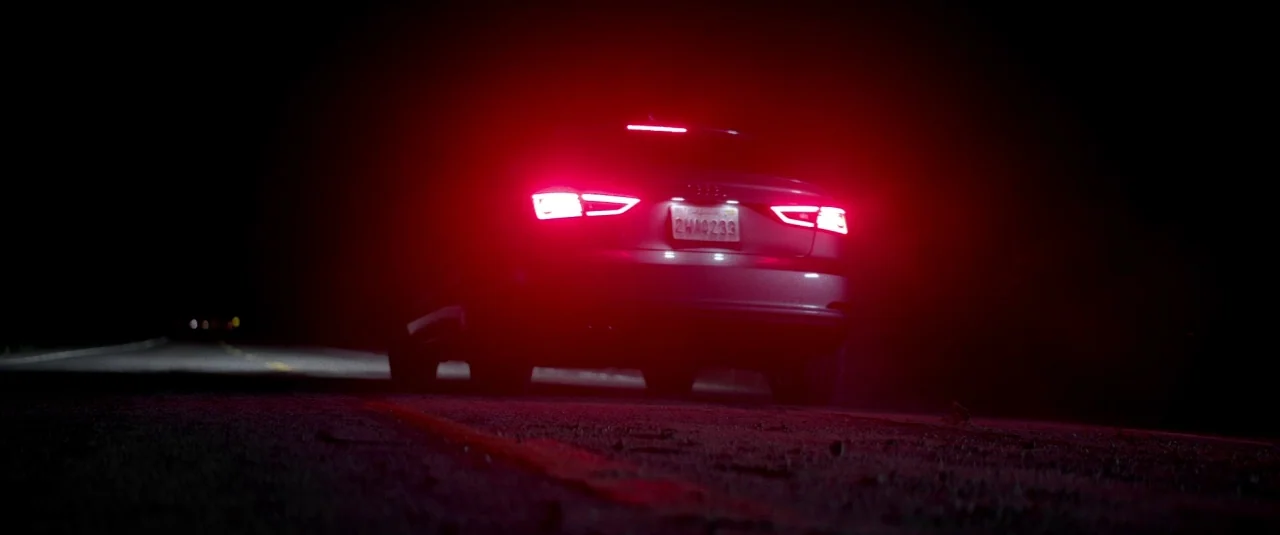 Southbound (2015) movie still: wide — A silver car is parked in the middle of a dark asphalt road at night. The bright red brake lights cast a glow…