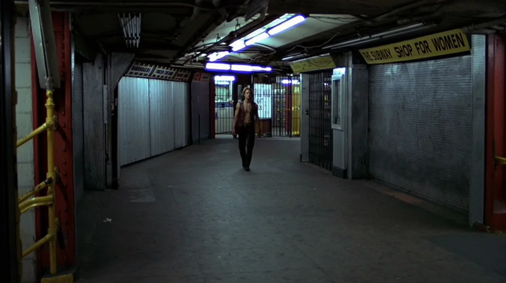 The Warriors (1979) movie still: wide — A dimly lit underground transit passageway features closed shop fronts with metal security shutters. A lone…