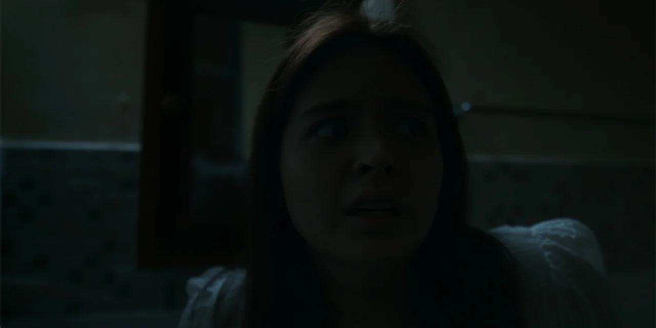 Nokturno (2024) movie still: closeup — A dimly lit room with tiled walls and blurred fixtures in the background. A woman in a white garment looks…