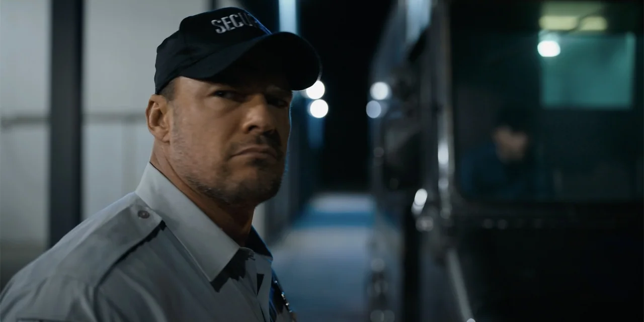 Playdate (2025) movie still: medium — A night exterior setting near a large truck on a paved surface. A male security guard in a uniform stands near…