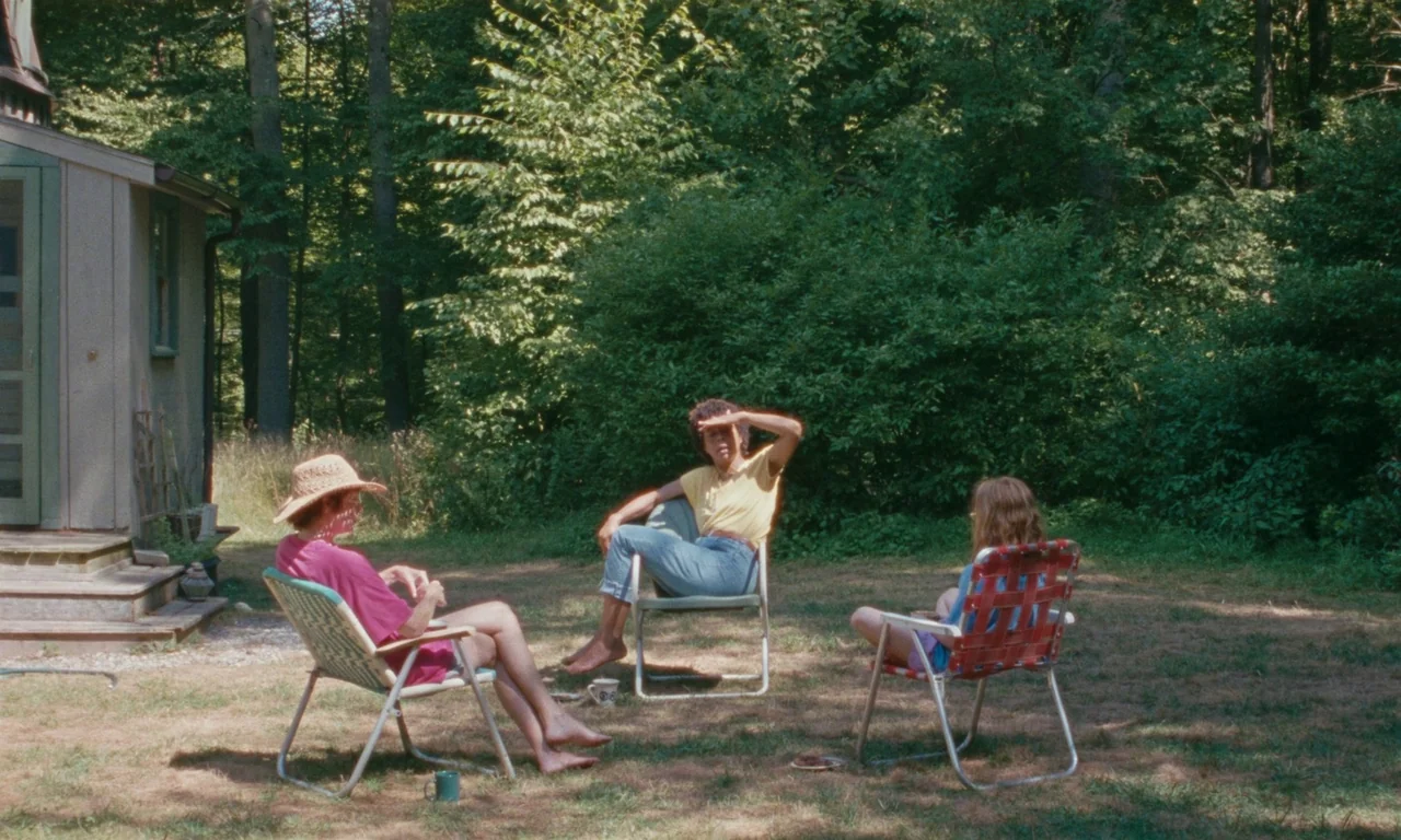 Janet Planet (2023) movie still: wide — A grassy clearing adjacent to a wooden cabin is surrounded by dense trees. Three young women sit in vintage…