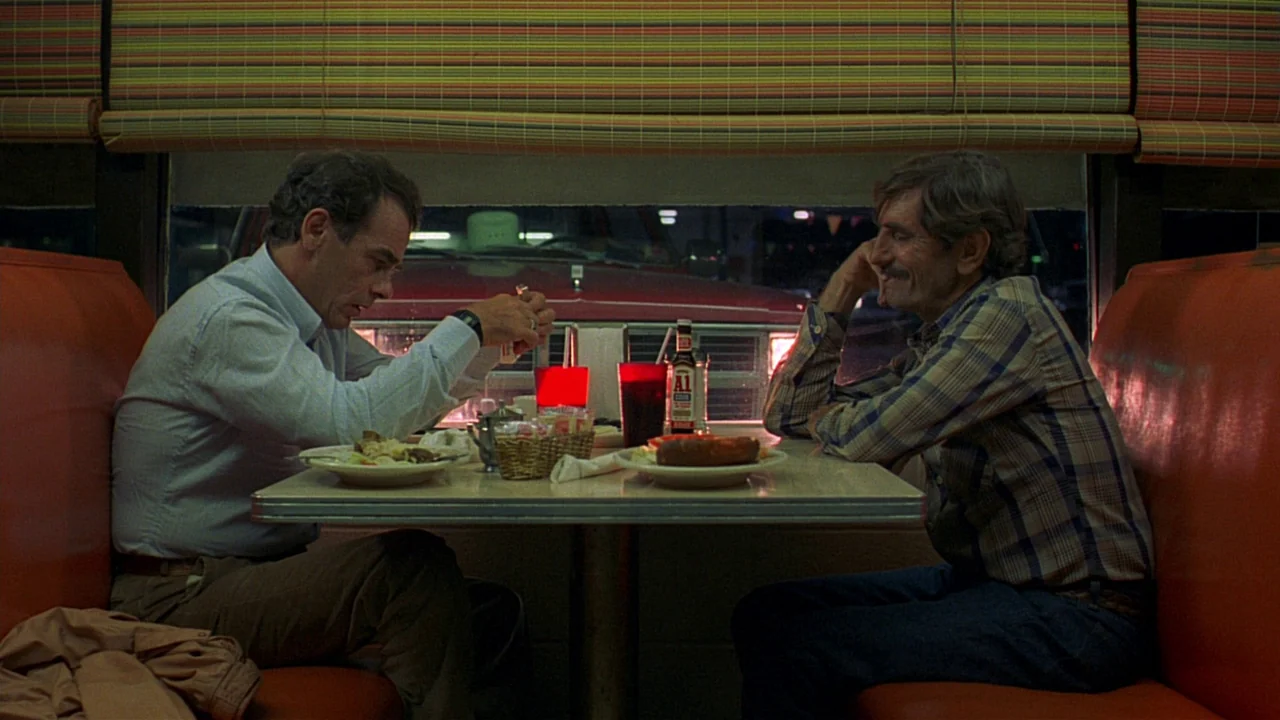 Paris, Texas (1984) movie still: medium — A diner booth with orange vinyl seats and a view of a parked car outside. Two men sit at the table in…