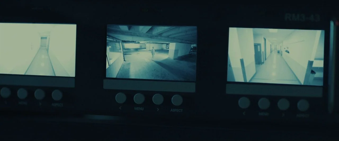 Come True (2020) movie still: wide — A close-up of a rack-mounted security console with three active surveillance monitors. Each screen shows a…