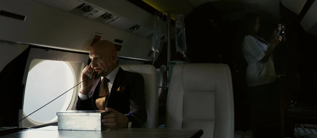 Self/less (2015) movie still: medium — The interior of a private jet cabin shows a man seated by a window. A medical IV drip bag hangs nearby while…