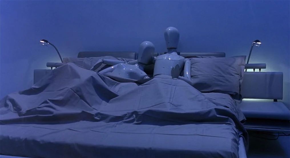 The Comfort of Strangers (1990) movie still: medium — A minimalist room contains a bed with two glossy white humanoid mannequin figures lying down.…