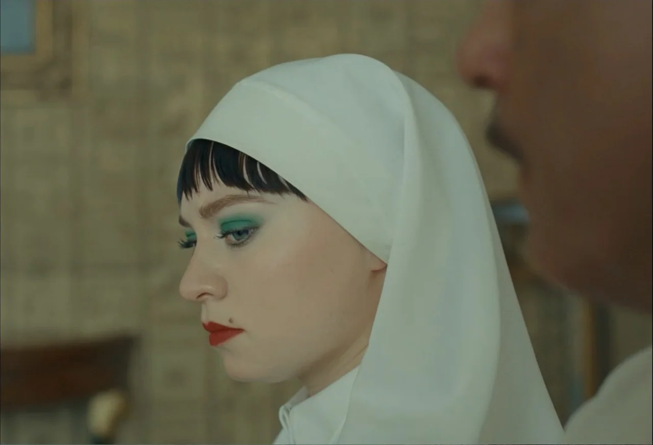 The Phoenician Scheme (2025) movie still: closeup — A light-toned room features patterned wallpaper and a blurred foreground figure. A woman wearing…